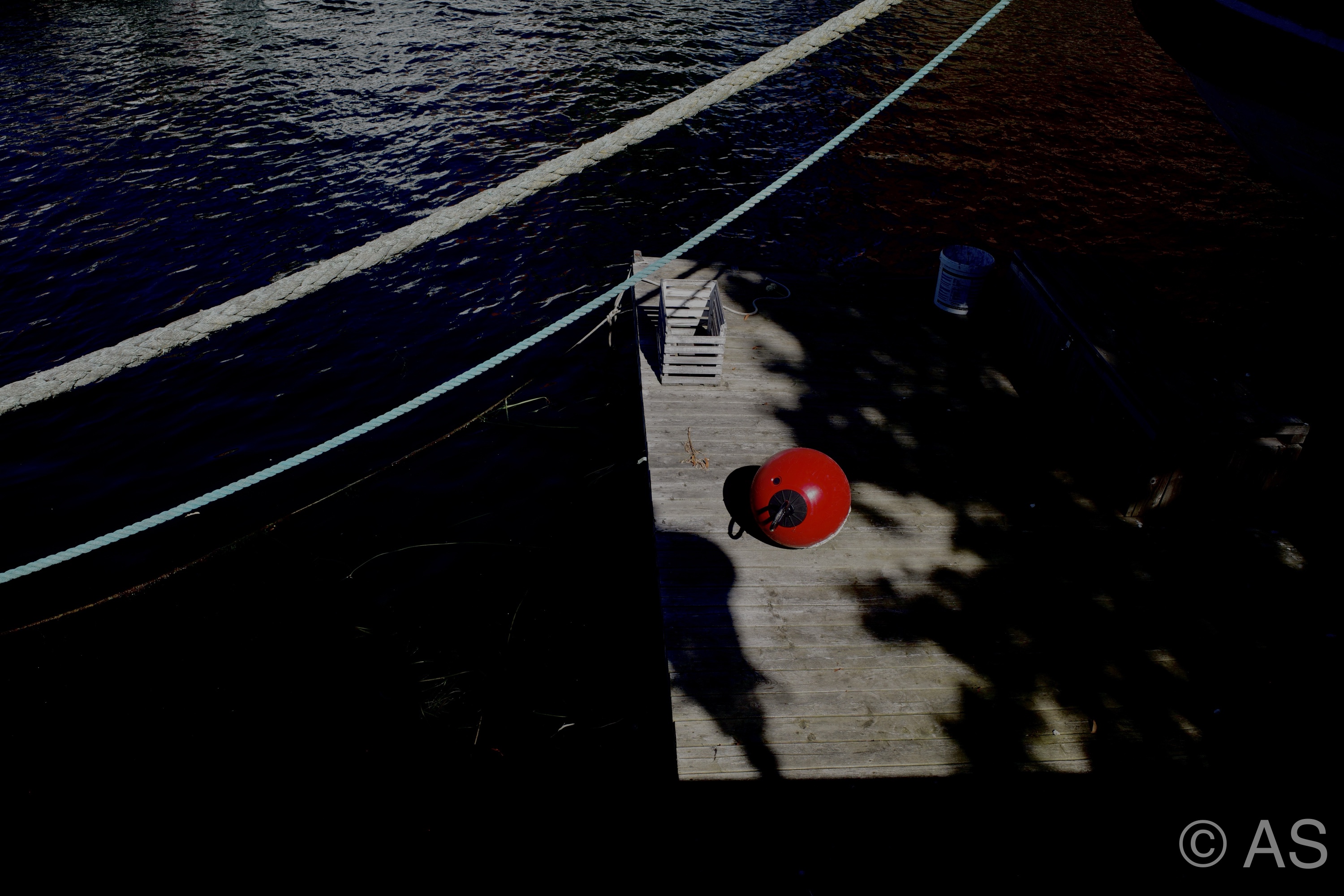 Wooden dock with a red buoy and boat hull from above, strong shadows across the planks